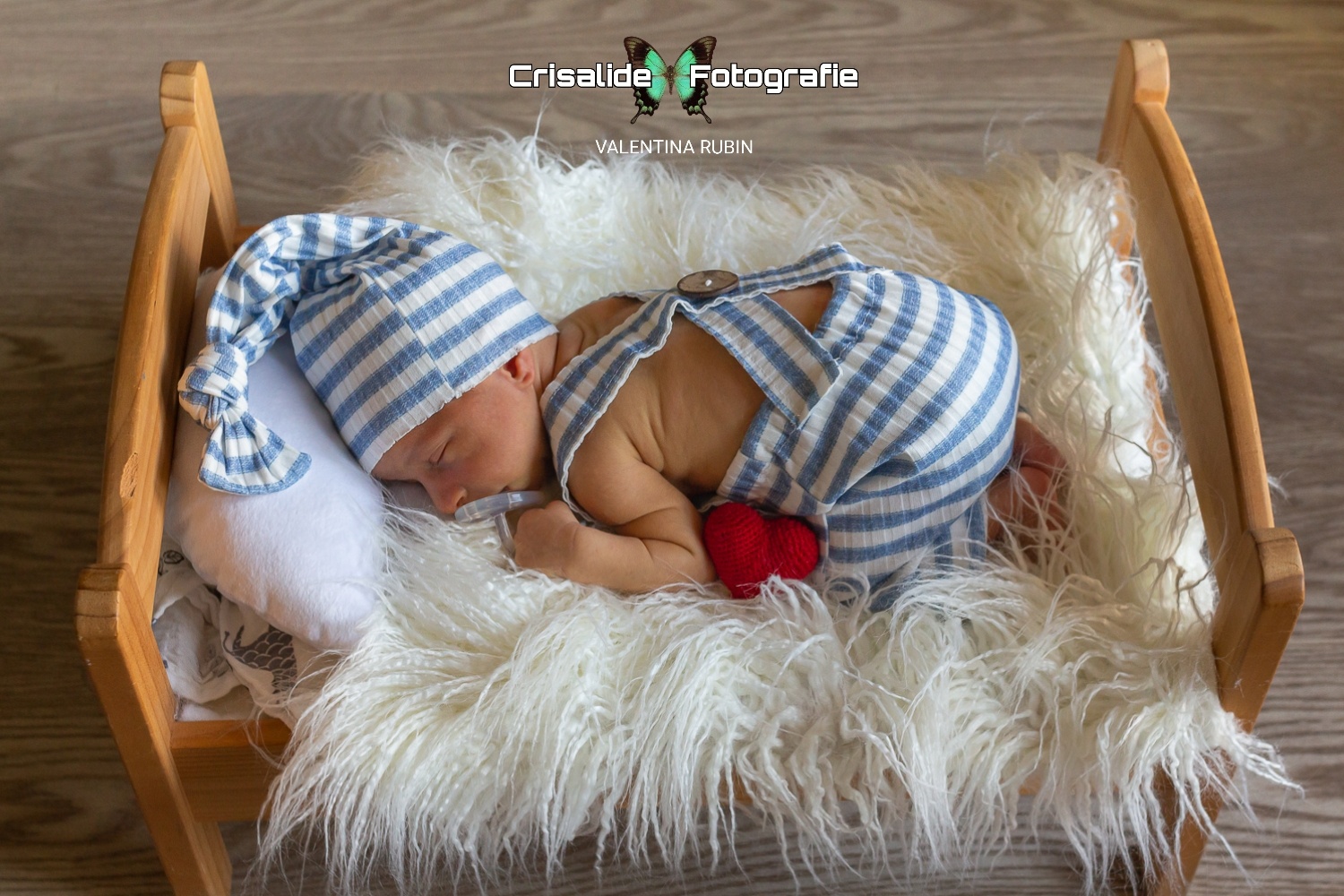Neonato disteso su un  lettino con salopette e berrettino coordinati a righe bianche e azzurre, cuore rosso al centro