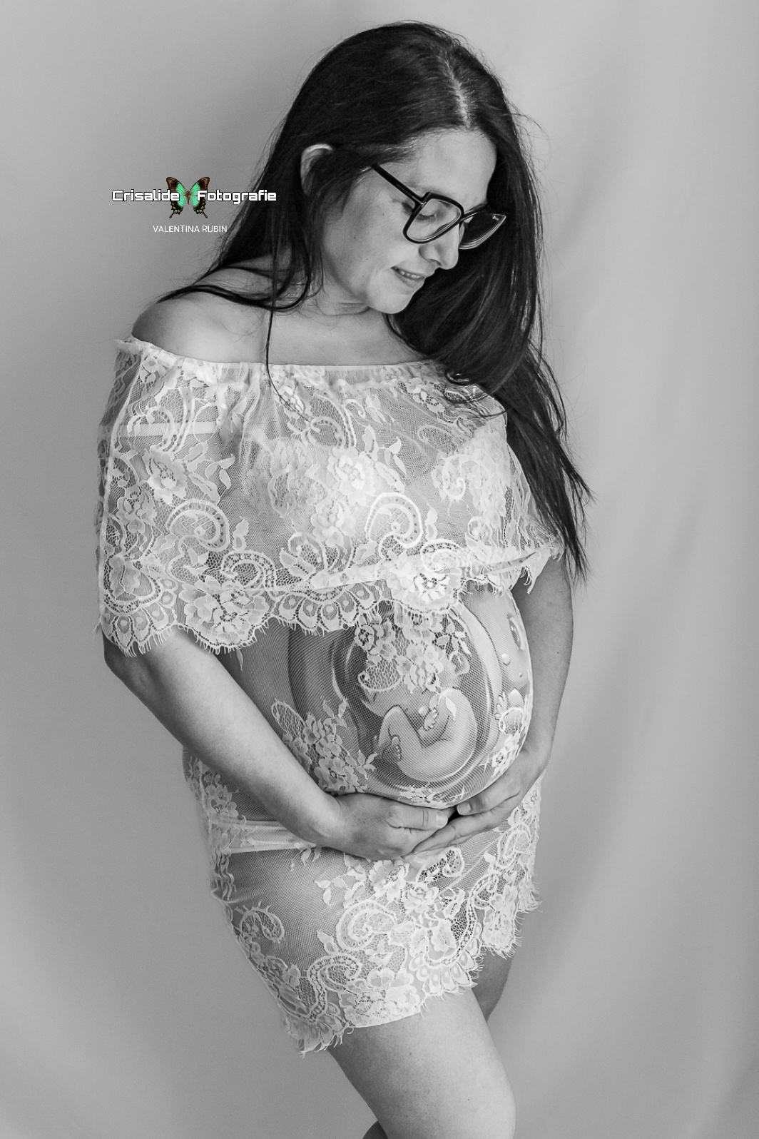 Mamma incinta in abito bianco di pizzo, tre quarti, sorriso verso il pancione, occhiali, gonna corta e belly painting che si intravede, gamba piegata su sfondo bianco