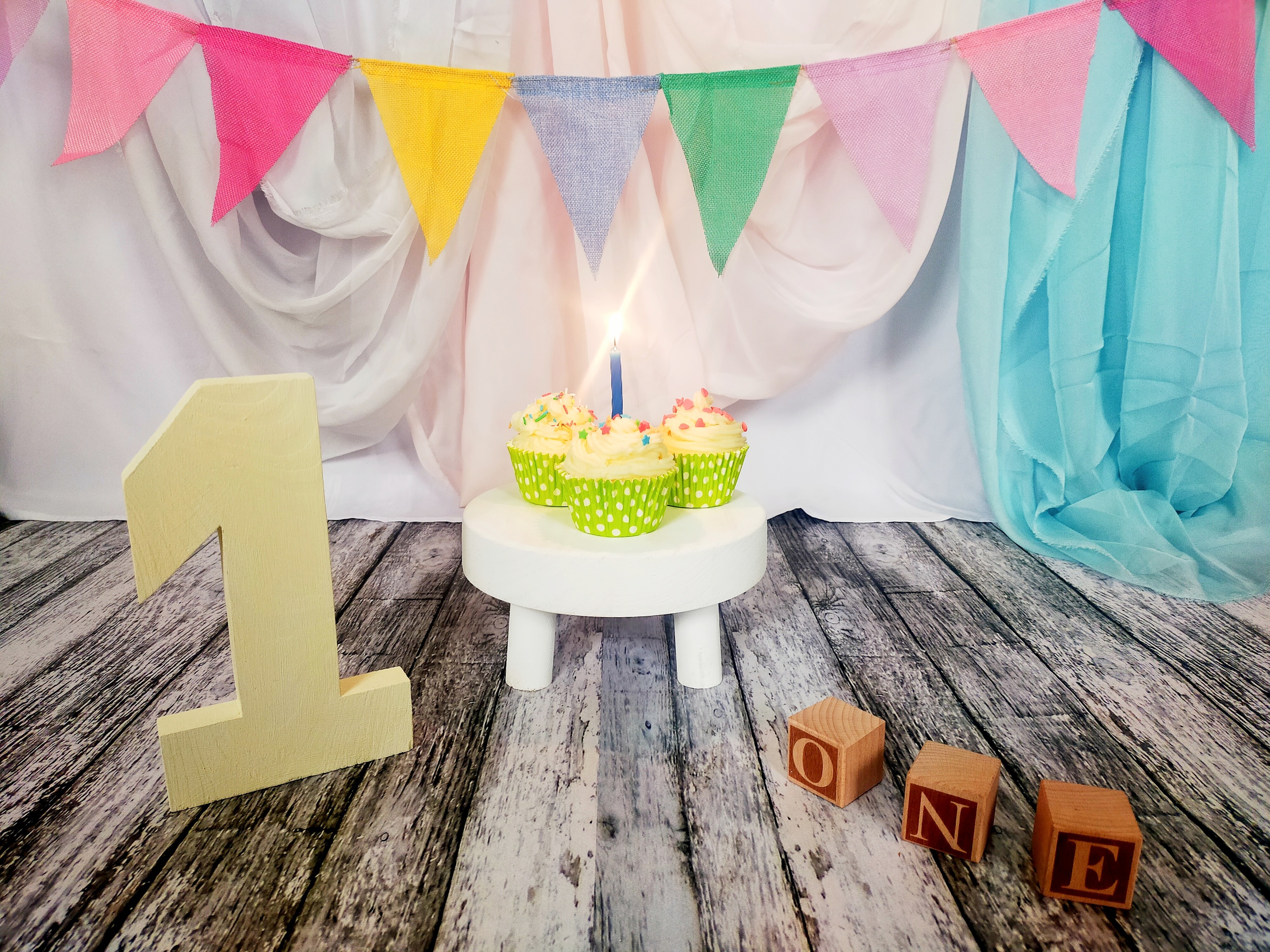 Set fotografico che comprende numero 1, cubi di legno con scritta one, alzatina per torte con sopra 3 cupcakes ed una candelina azzurra accesa. Lo sfondo è colorato con bandierine