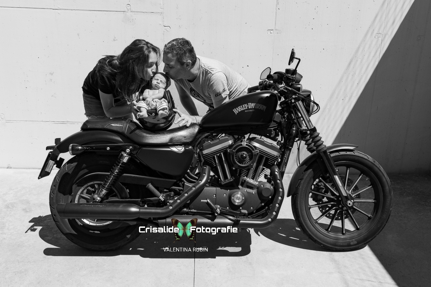 Mamma e papà baciano la testina del neonato che dorme dentro un casco appoggiato su una moto Harley Davidson, scatto newborn in bianco e nero