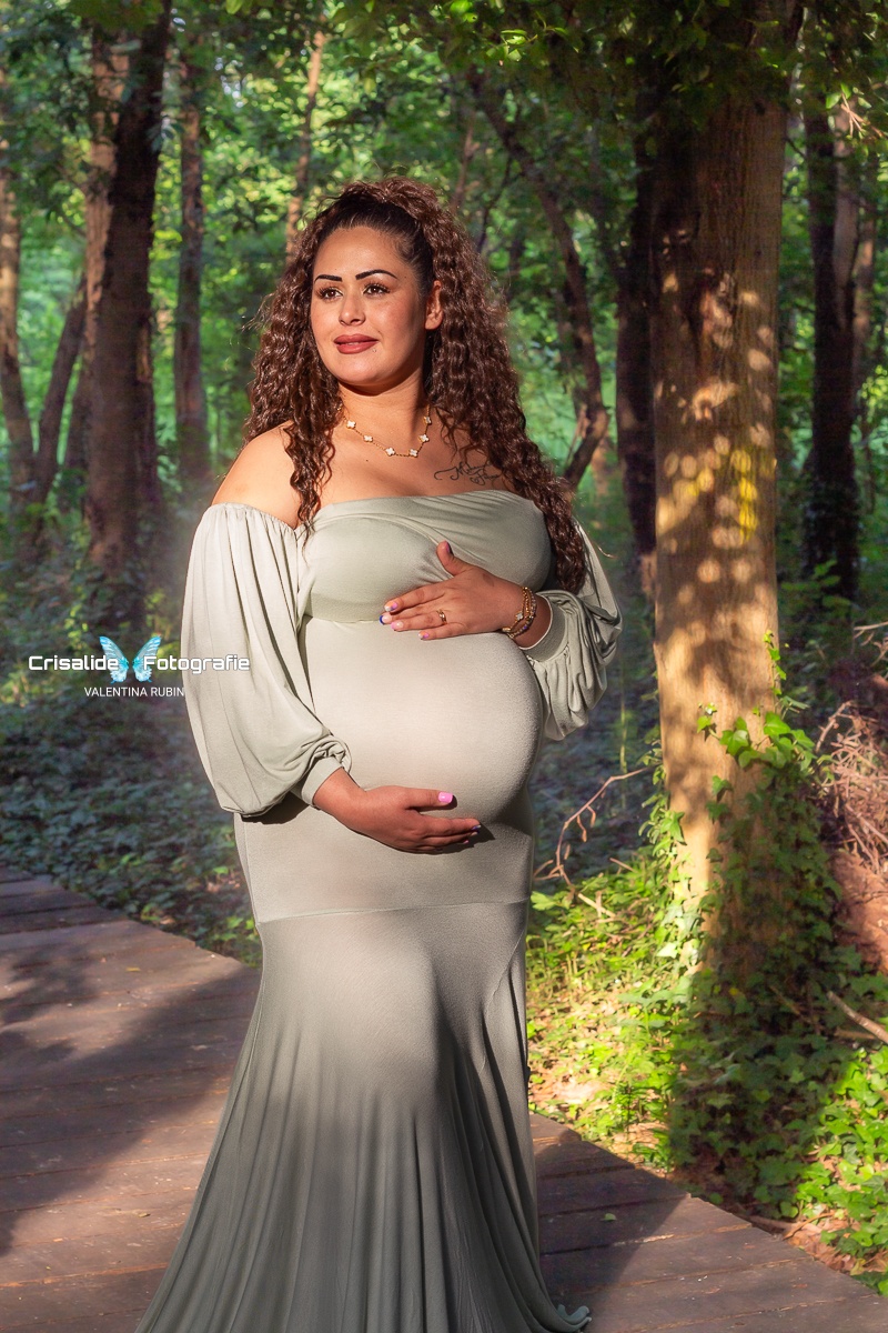 Donna incinta con abito verde chiaro a sirena, ritratta in verticale tra gli alberi. È girata di tre quarti, una mano sopra e una sotto la pancia, sorride guardando di lato. Capelli mori ondulati sulle spalle, illuminata dalla luce naturale filtrata dal verde. Scatto di maternità naturale ed emozionale