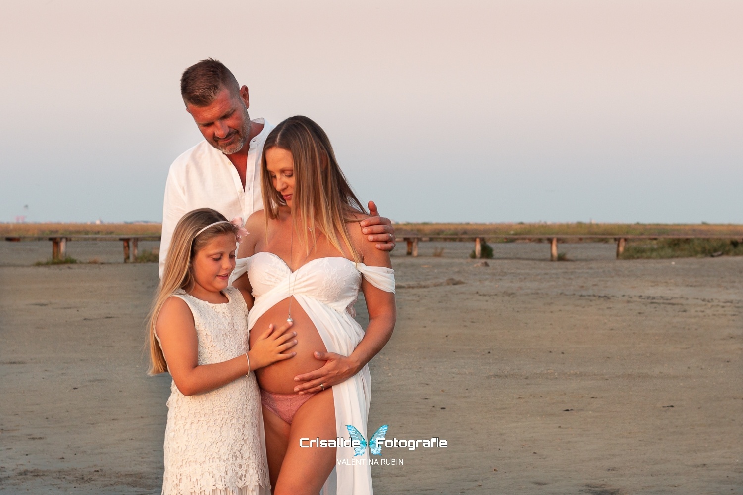 Mamma incinta in abito bianco in chiffon con spalle scoperte sulla spiaggia, con la figlia accanto in abito bianco e cerchietto tra i capelli biondi, e il papà alle spalle con camicia bianca e barba brizzolata. La mamma guarda la figlia, la figlia guarda la pancia, il papà poggia la mano sulla spalla della mamma. Mamma e figlia con mani sulla pancia. Sfondo spiaggia con pontile