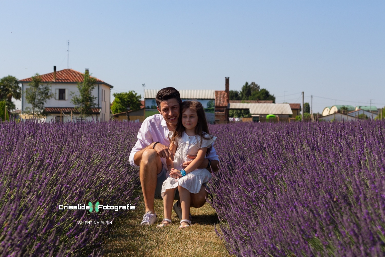 Papà accucciato in camicia bianca che tiene seduta la figlia vestita di bianco in un campo di lavanda sotto il cielo azzurro