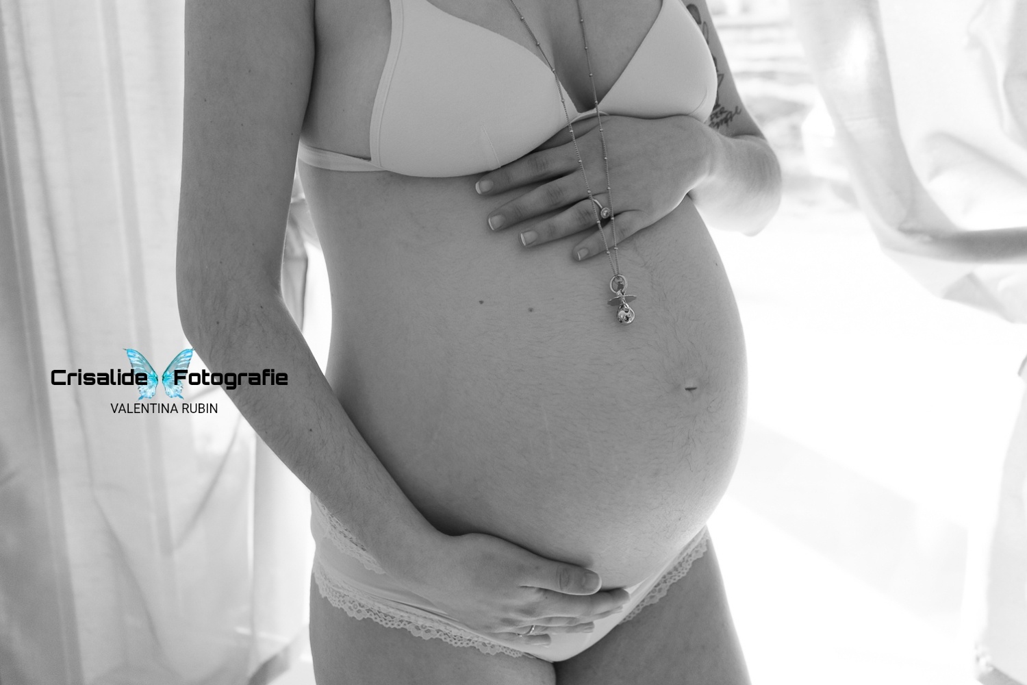 Scatto in bianco e nero. Primo piano del pancione girato di tre quarti, una mano sopra e una sotto, finestra con tenda sullo sfondo