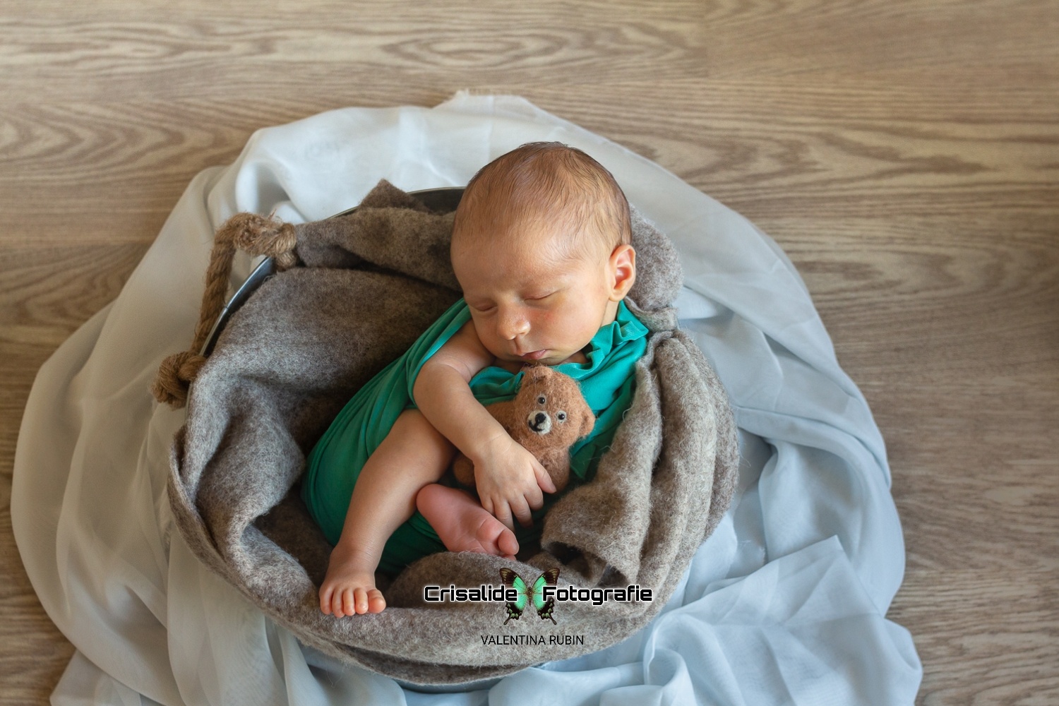 Neonato che dorme fasciato di verde all'interno di un secchio con coperte, orsacchiotto e chiffon in azzurro, su pavimento in legno