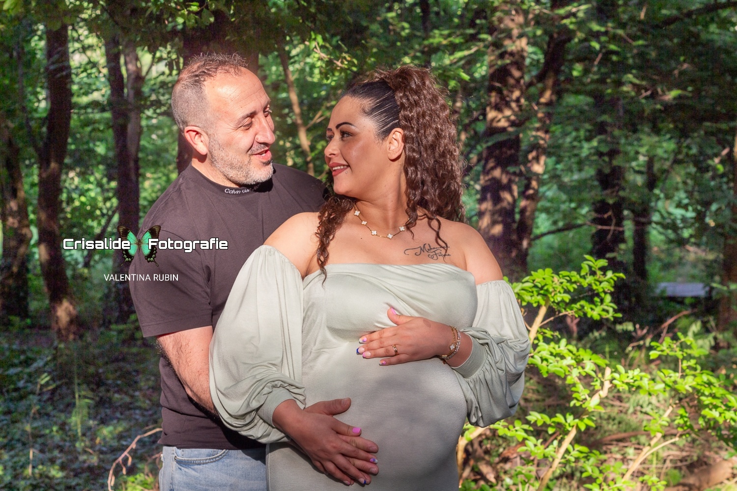 Mamma incinta vestita in abito verde chiaro a sirena, maniche lunghe e spalle scoperte, viso girato verso sinistra sorridente che guarda il marito. Una mano sopra e una sotto la pancia, mano del marito sopra la sua. Marito dietro di lei a sinistra, maglia nera e jeans, sorriso rivolto a lei. Illuminati da luce solare filtrata dagli alberi, sfondo naturale con alberi e vegetazione