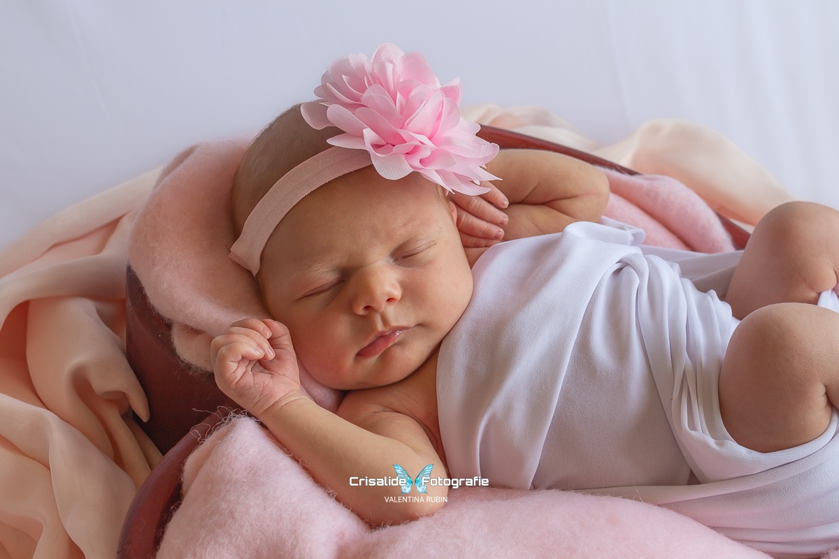 Neonata che dorme in una cestina a forma di cuore, fasciata di bianco, con fiore rosa sulla testa e tessuti rosa su sfondo bianco