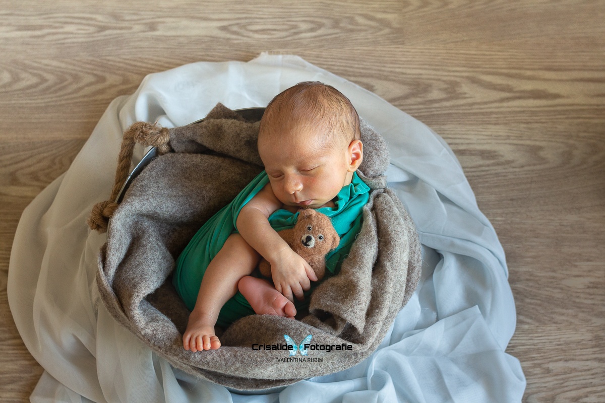 Neonato che dorme fasciato di verde all'interno di un secchio con coperte, orsacchiotto e chiffon in azzurro, su pavimento in legno