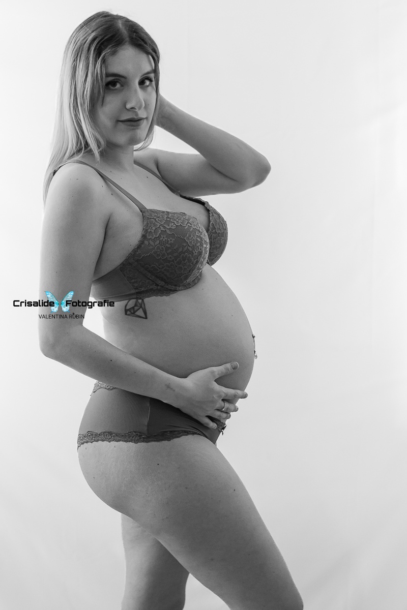 Mamma incinta di profilo in intimo scuro, gamba piegata, una mano sotto la pancia, e una dietro la testa, sguardo in camera, luce laterale su sfondo bianco